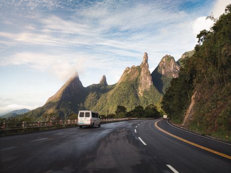 Van traveling on a scenic road towards the iconic Dedo de Deus mountain under a bright sky.