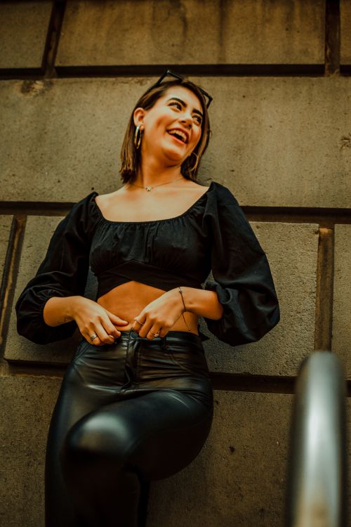Free Happy woman in black crop top and leather pants leaning against urban wall, smiling. Stock Photo