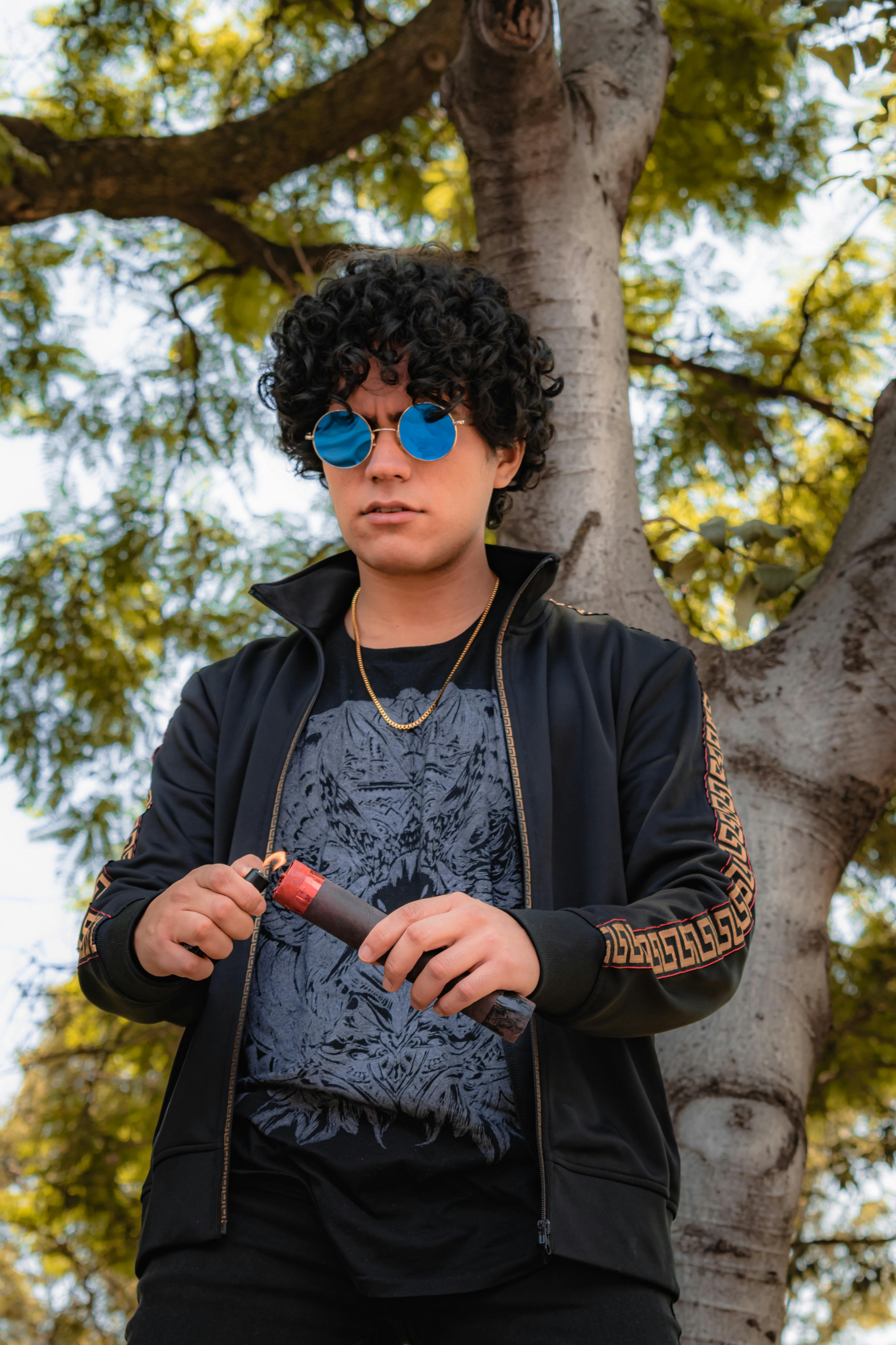 A stylish young adult in a black jacket holding a smoke bomb stands under a tree.