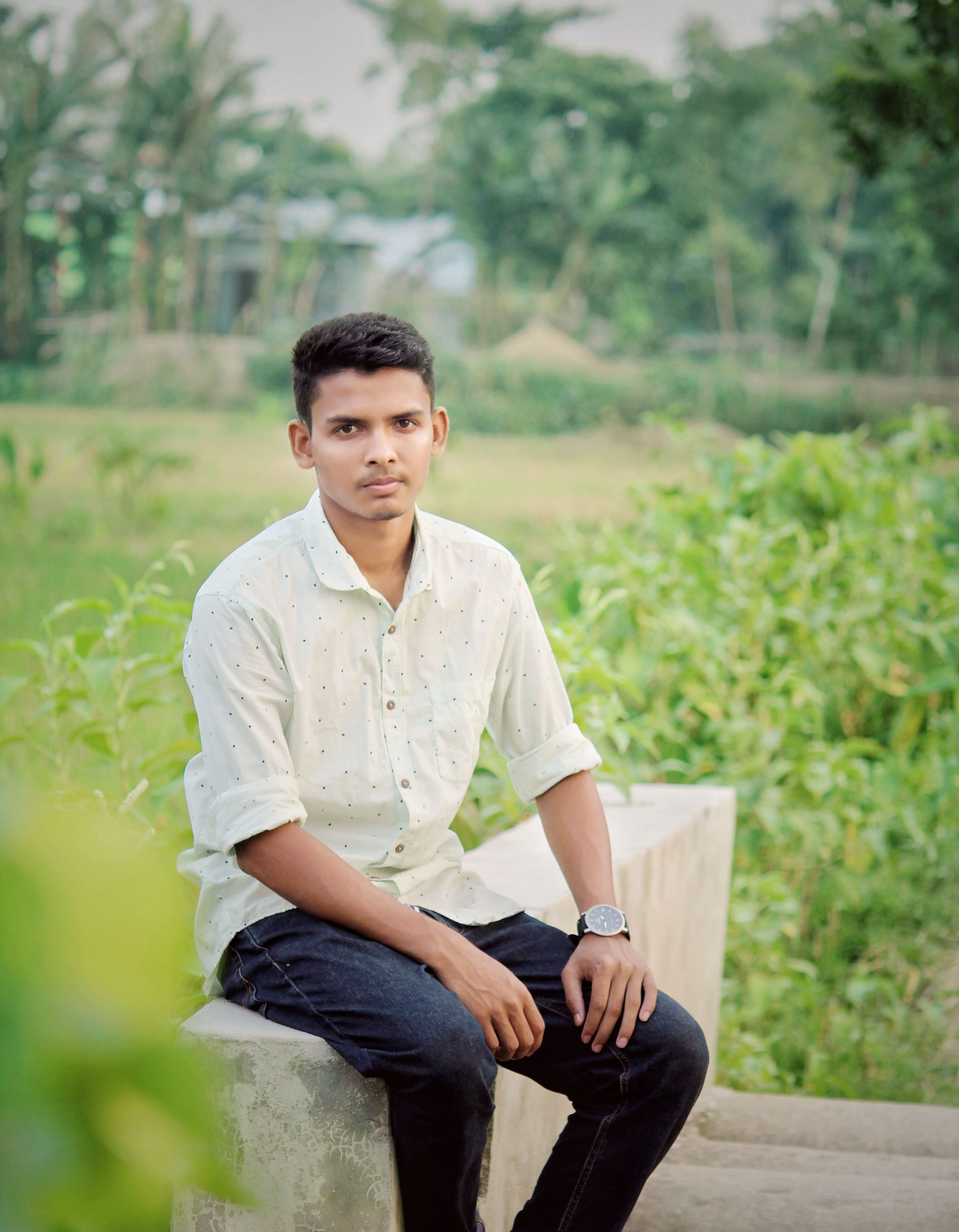 Asian man sitting on concrete bench in lush green outdoor setting, casual white shirt.