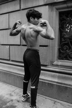 A shirtless man flexes his muscles against an urban background in a monochrome photo.