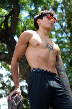 Shirtless man with tattoos and sunglasses standing outdoors against a tree.