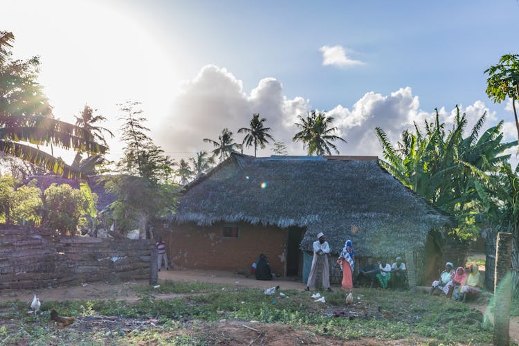 People Sitting And Standing Beside Nipa Hut House In Village Surrounded With Banana Trees