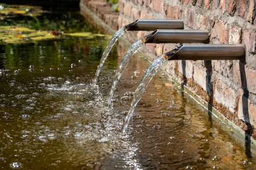 Free Crystal clear water streams from a modern stainless steel fountain into a serene outdoor pond. Stock Photo