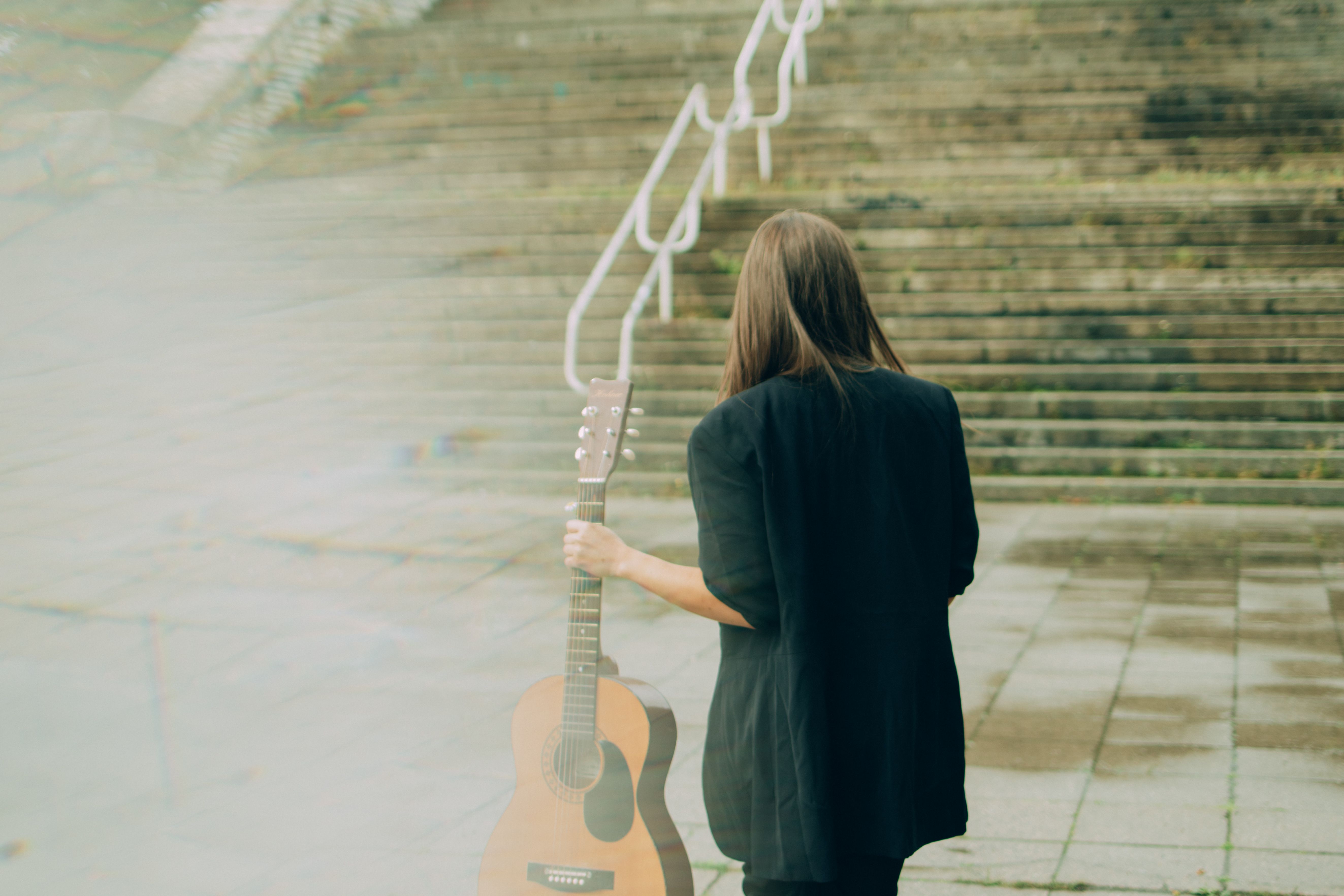 A woman stands outdoors holding an acoustic guitar near a concrete staircase, seen from the back.