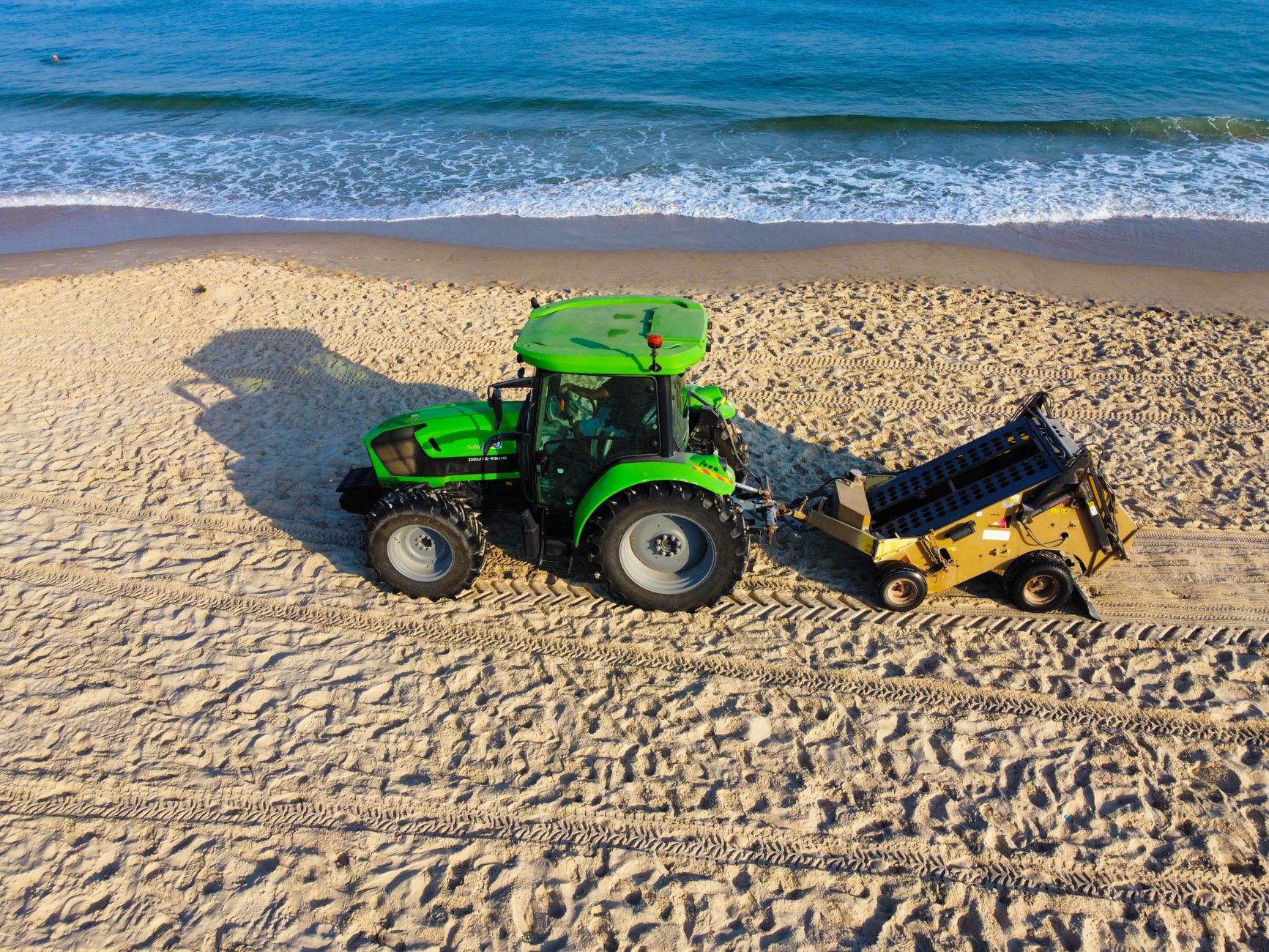Aerial view of a green tractor cleaning a sandy beach near the ocean waves on a sunny day.