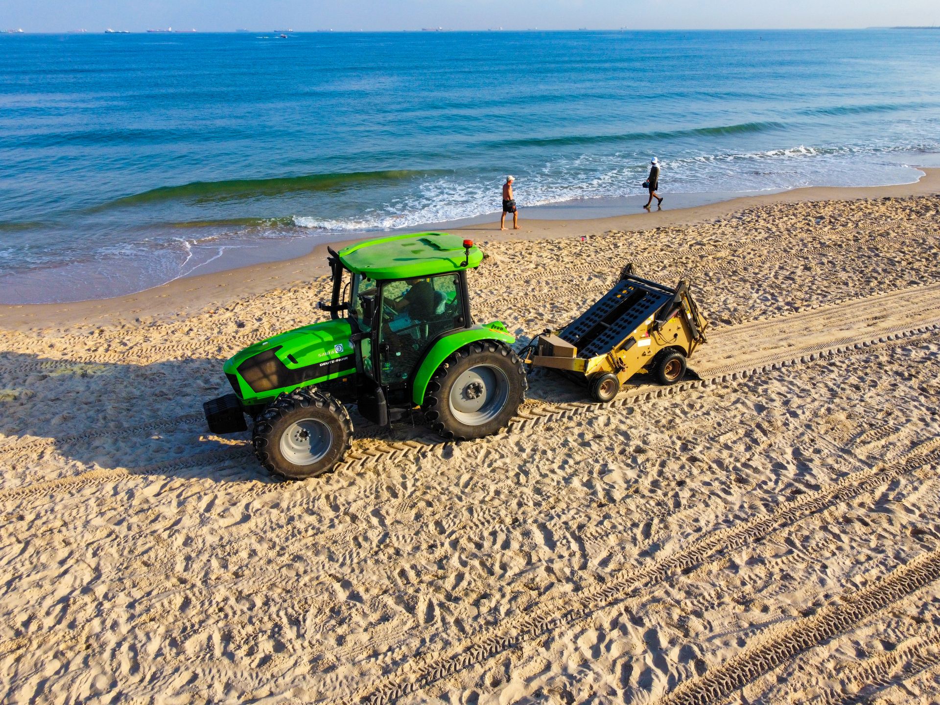 Green tractor with attachment cleaning the sandy beach while people walk along the shoreline.
