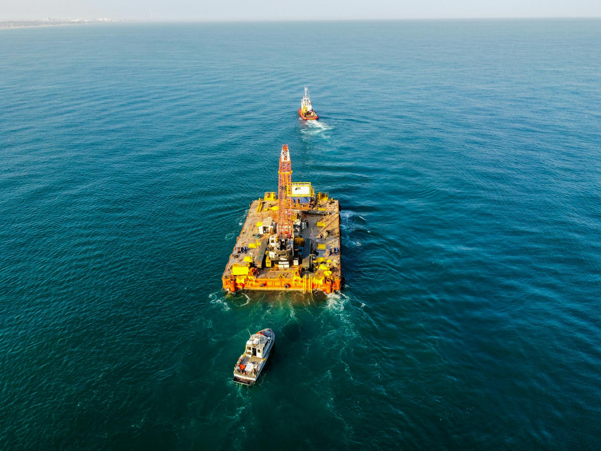 Aerial shot of heavy equipment on a barge in the sea, supported by tugboats.