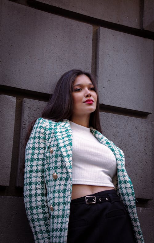 A Woman in White Tank Top and Green Plaid Coat Standing Beside the Concrete Wall