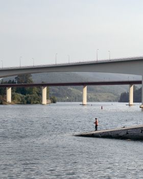 A man fishes in a river under a bridge in Penafiel, Portugal during summer.