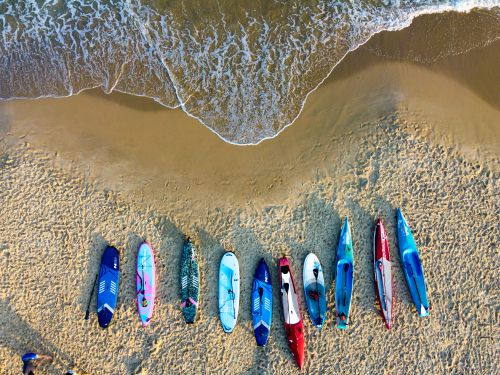Free Aerial view of colorful kayaks and paddleboards lined up on a sandy beach with gentle waves. Stock Photo