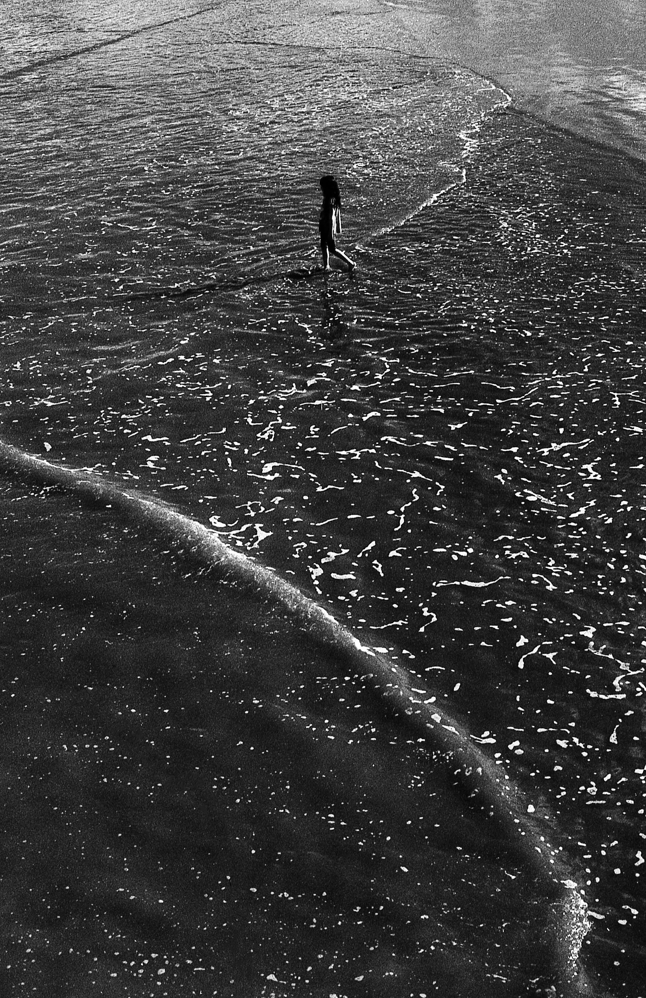 A lone child walks along the shoreline in this monochrome beach photo.