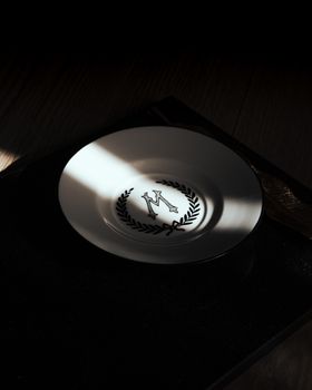 A sophisticated white plate featuring a monogram 'M' with a laurel wreath, presented in a dramatic light setting.
