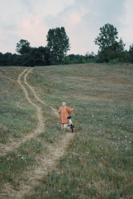 A person in an orange coat walking a bicycle along a rural pathway through a grassy field.