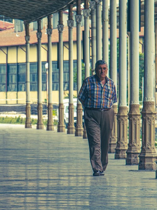 A Man in Blue and White Plaid Shirt Walking Near Columns
