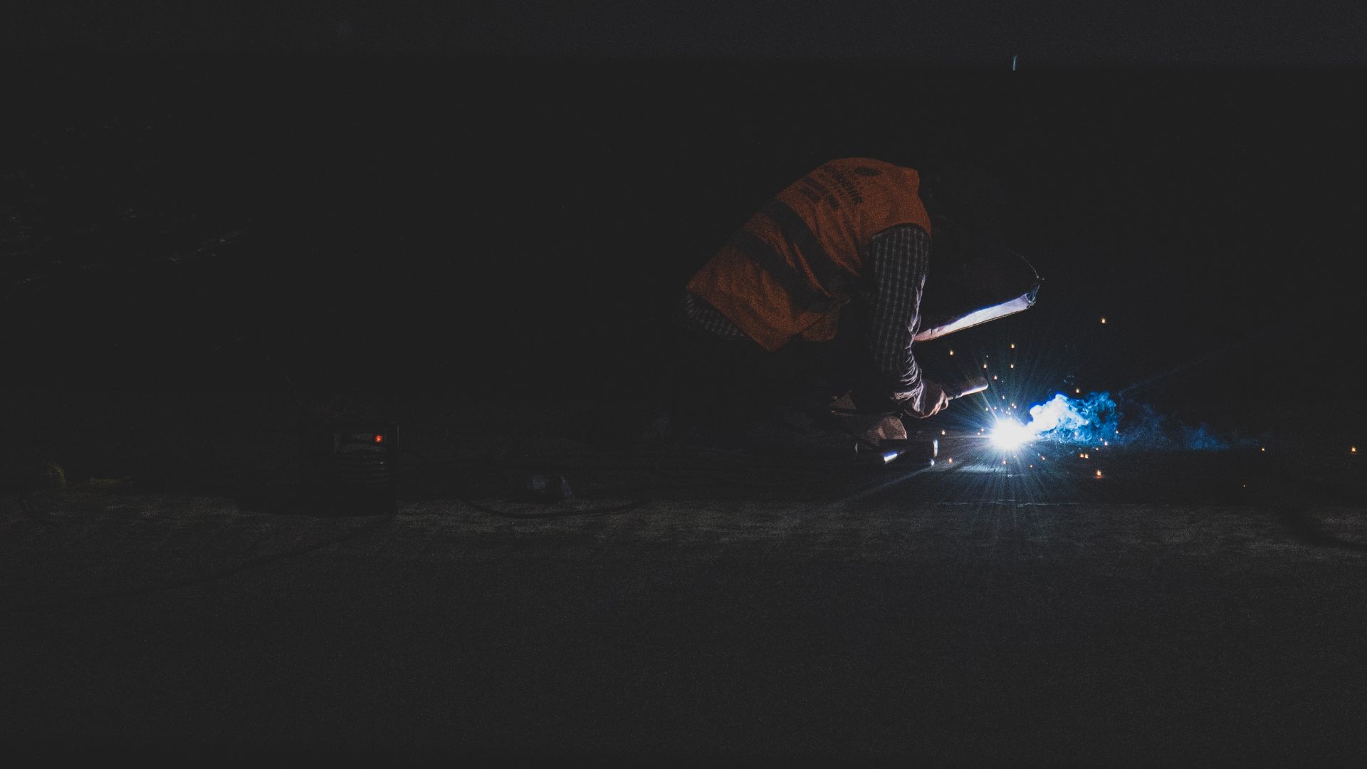 A welder creates sparks in dark surroundings wearing safety gear at night.