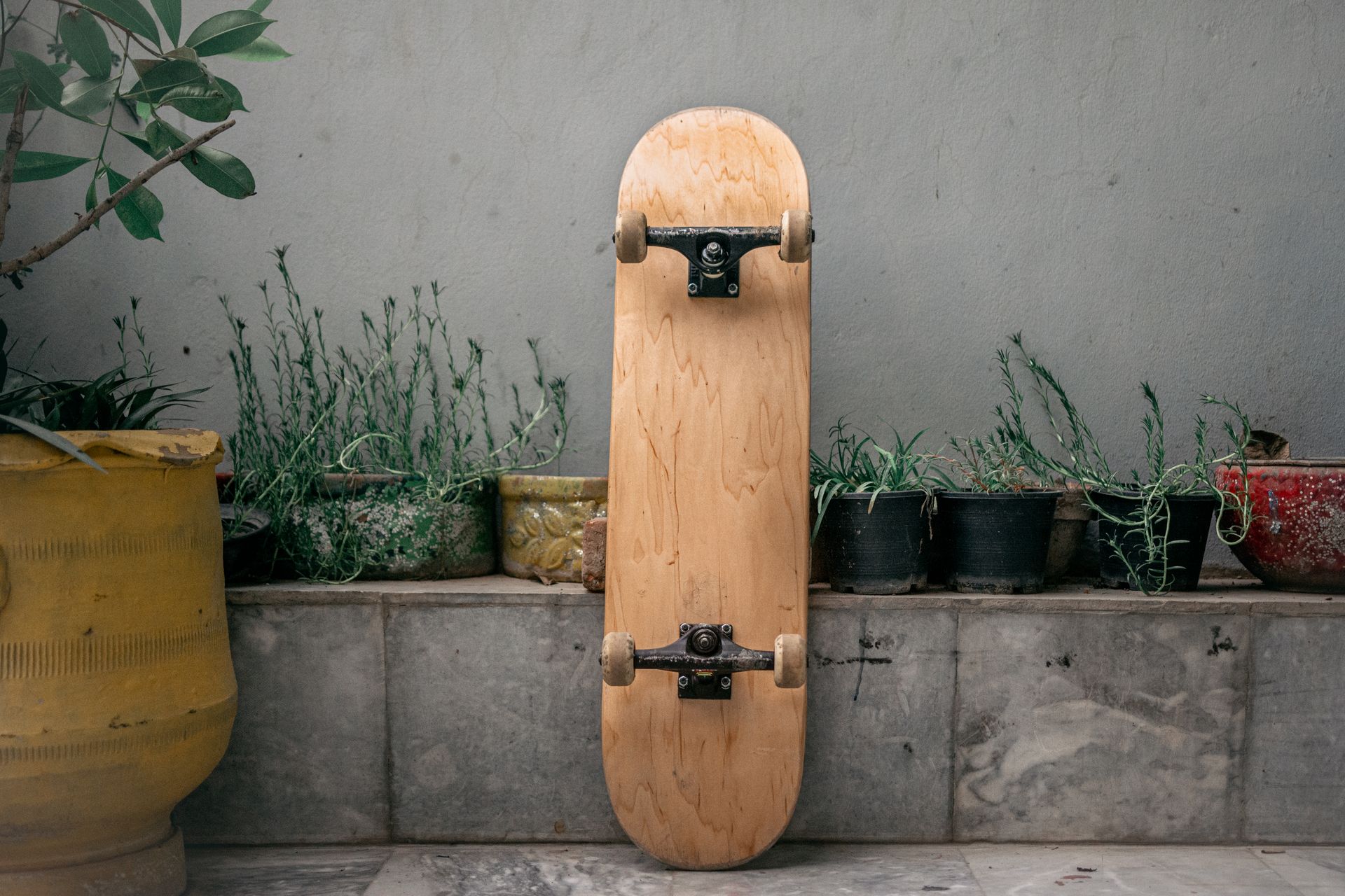 Natural wooden skateboard leaning against a rustic outdoor planter setup.