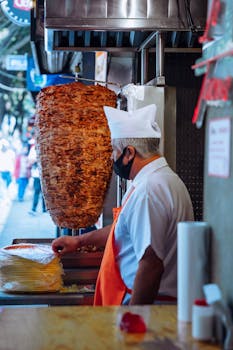 Chef in white uniform and face mask prepares shawarma kebabs at an outdoor street food stand.