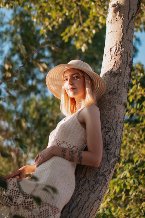 Woman in Beige Sleeveless Dress Wearing Sun Hat Leaning on Tree Trunk