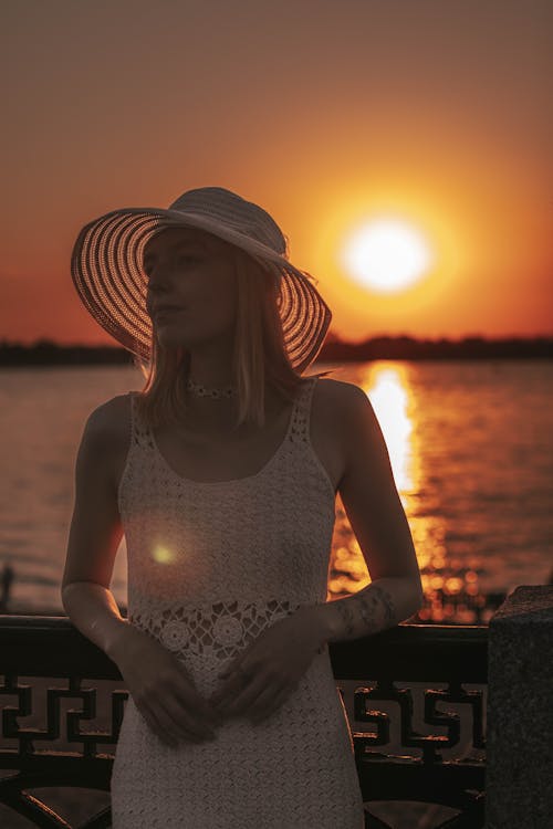 Woman in Brown Sun Hat and White Crochet Dress Leaning Against Hand Rail During Golden Hour