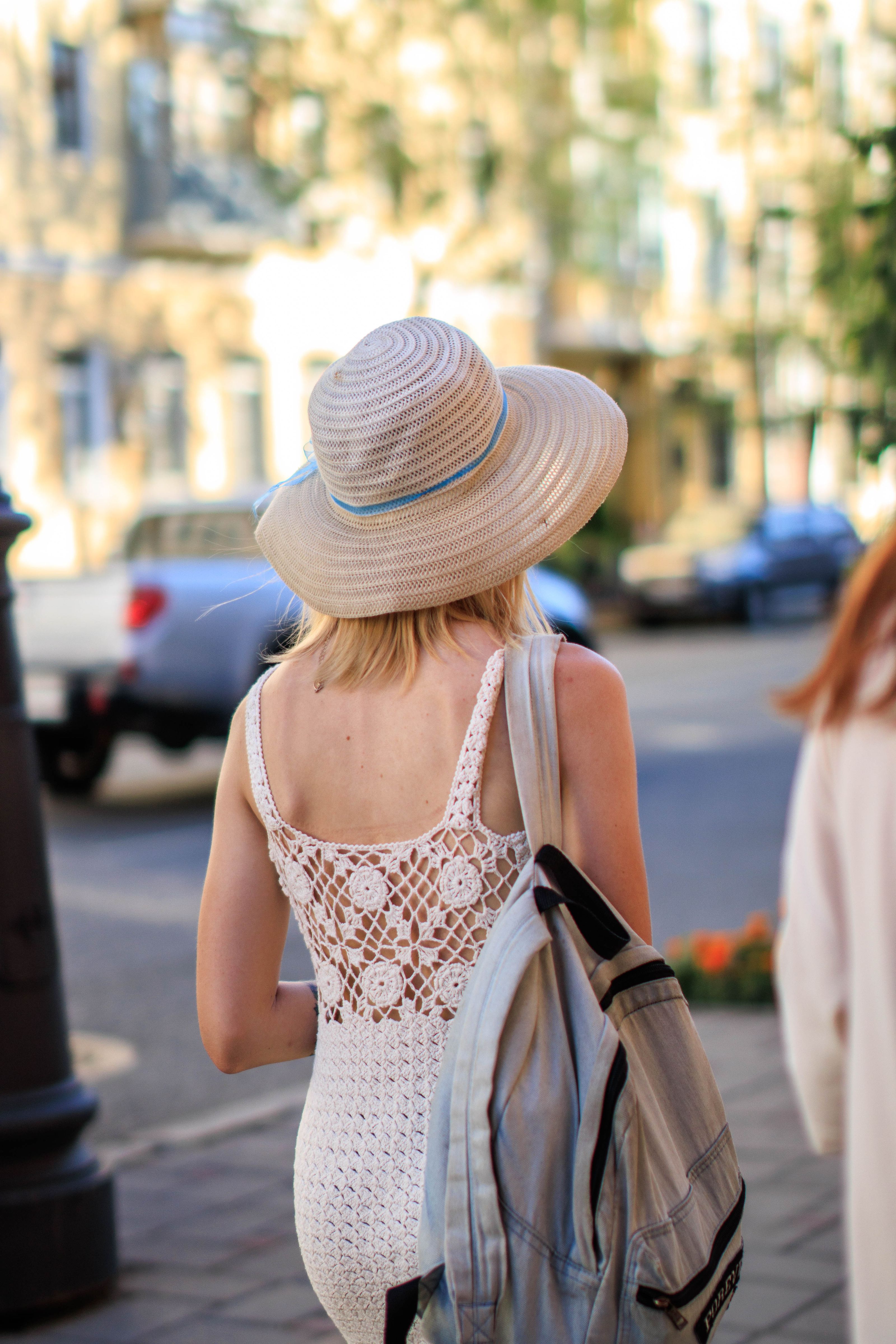 Uma mulher estilosa com chapéu de sol e vestido de crochê carrega uma bolsa enquanto passeia por uma rua ensolarada da cidade.