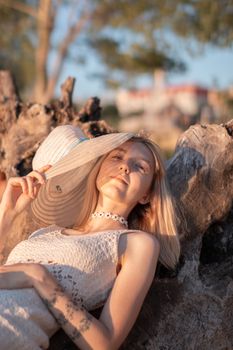 A woman reclines on a tree stump outdoors, wearing a sun hat and white dress.