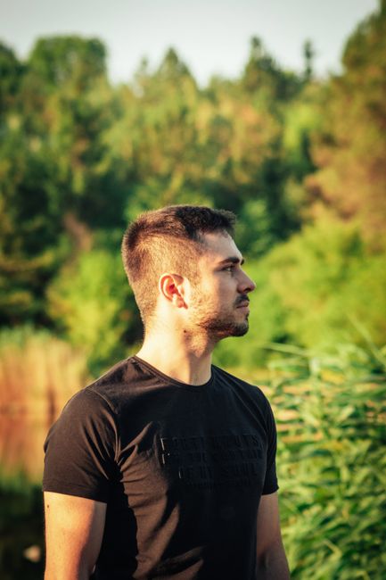 Portrait of a man in a black T-shirt looking aside amid lush greenery, under natural light.