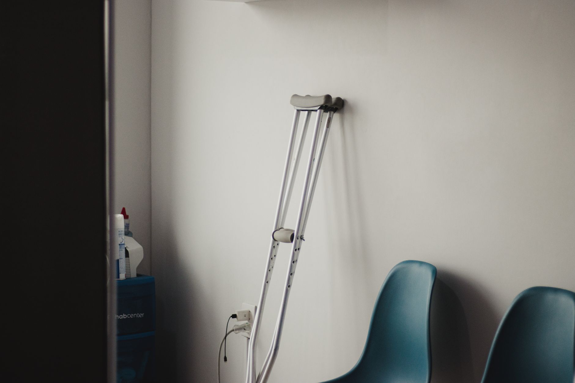 Crutches resting against a wall in a minimalist waiting room with blue chairs.