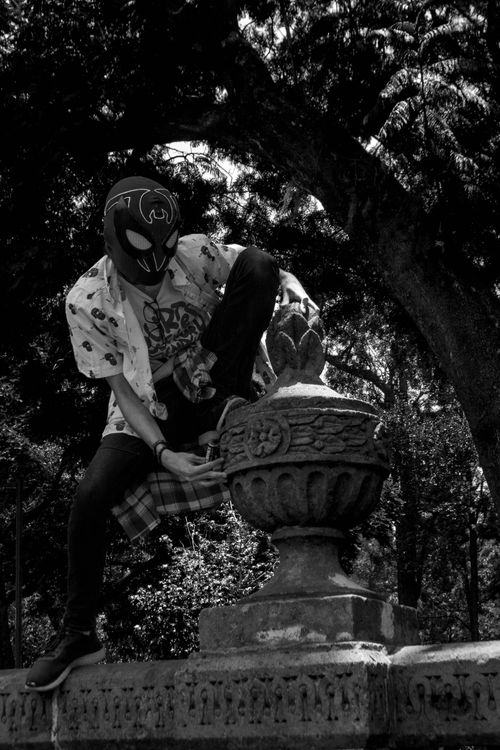 Free A masked person sitting on a stone structure under trees in monochrome. Stock Photo