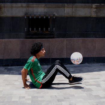 A man performing freestyle tricks with a soccer ball on a city sidewalk.
