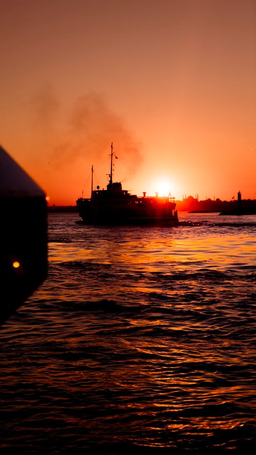 Free A silhouetted ship glides across the ocean at sunset, with a golden sky and serene waters. Stock Photo