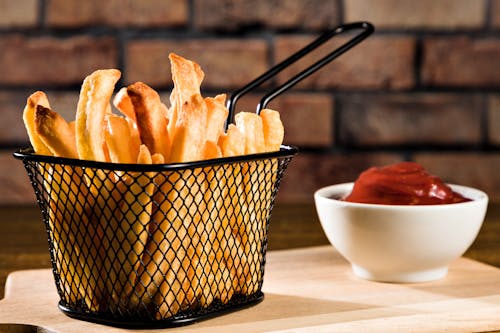 Closeup of a Frying Basket with French Fries and Bowl of Ketchup against Brick Wall