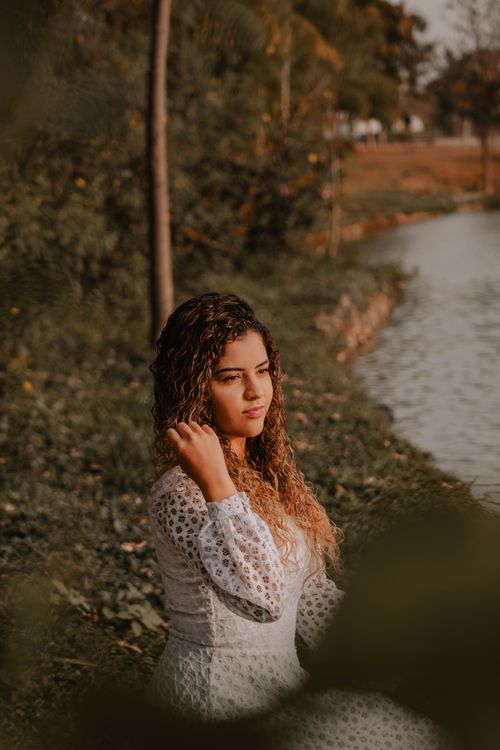 Free Woman in a white dress with curly hair sitting by a tranquil riverside at sunset. Stock Photo