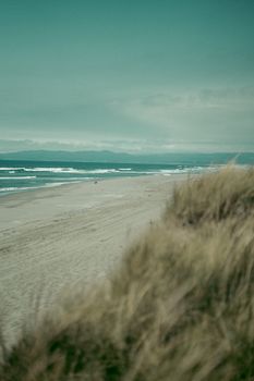 Peaceful beach scene with waves, sandy shore, and grassy dunes under a cloudy sky.