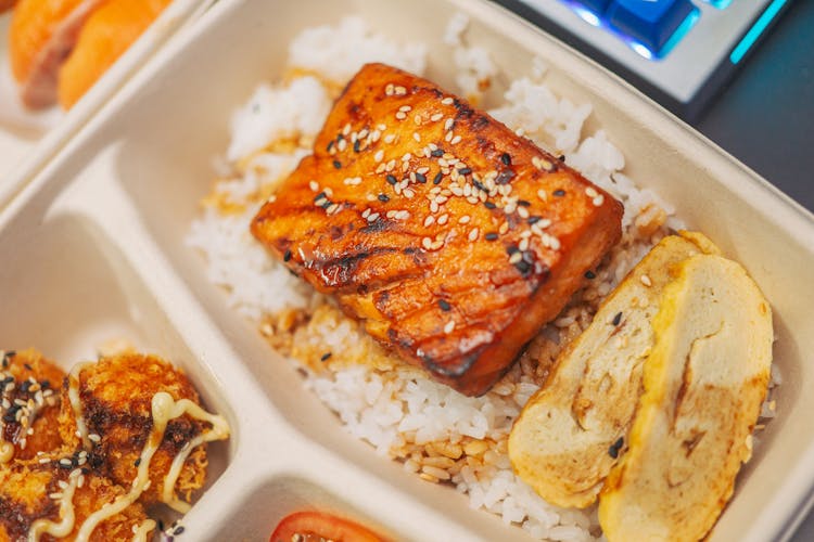 A Rice Topping Of Roasted Salmon With Sesame Seeds And Scrambled Egg In A Take Out Container