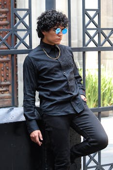 Stylish young man in black attire leaning against a metal gate outdoors.