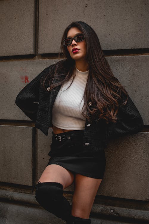 Woman in Black Cardigan and White Crop Top and Black Skirt Sitting on Concrete Wall