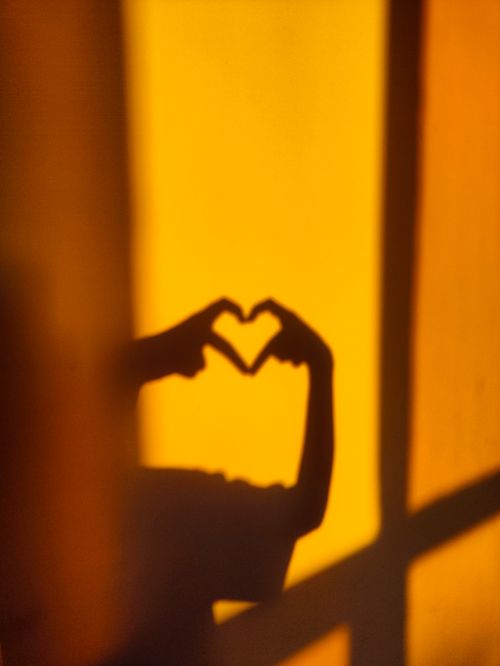 Free Silhouette of hands forming a heart shape against a warm yellow wall. Stock Photo