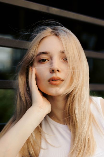 Close-up portrait of a blonde woman with long hair posing outdoors, hand on cheek.