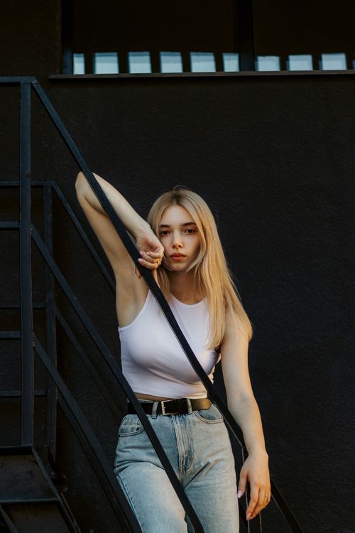 Free A confident young woman with long blonde hair poses on a black metal staircase in a modern, urban setting. Stock Photo