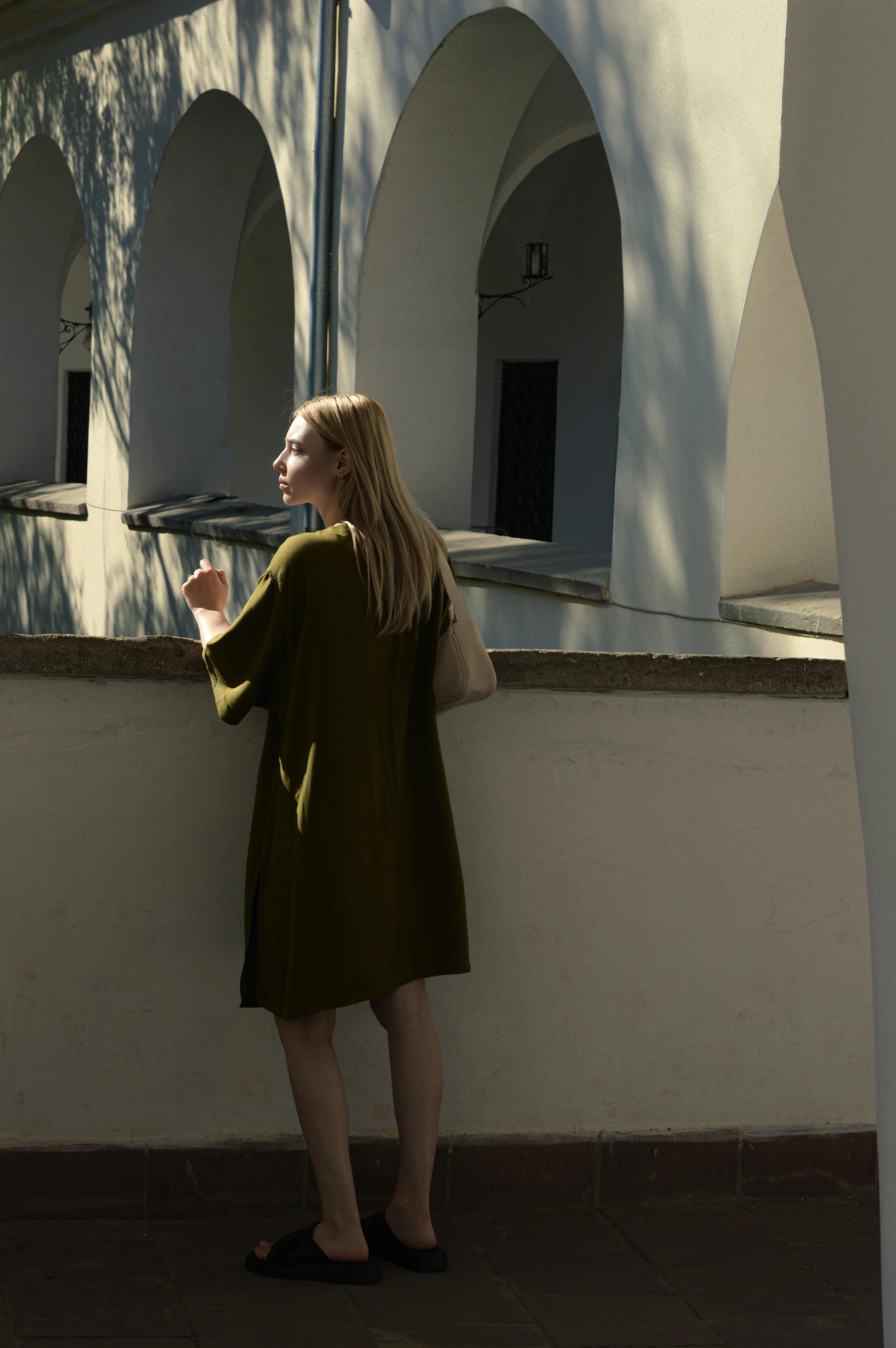 A woman stands on a balcony, surrounded by arches, in warm sunlight.