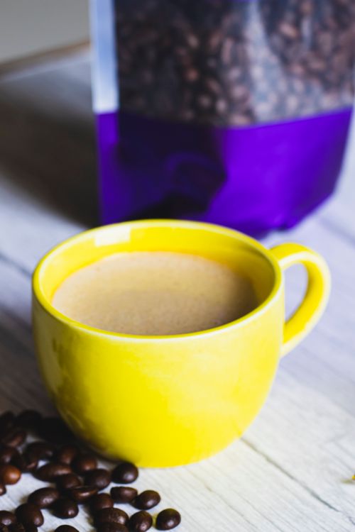 Free A warm beverage in a yellow cup surrounded by coffee beans on a wooden table. Stock Photo