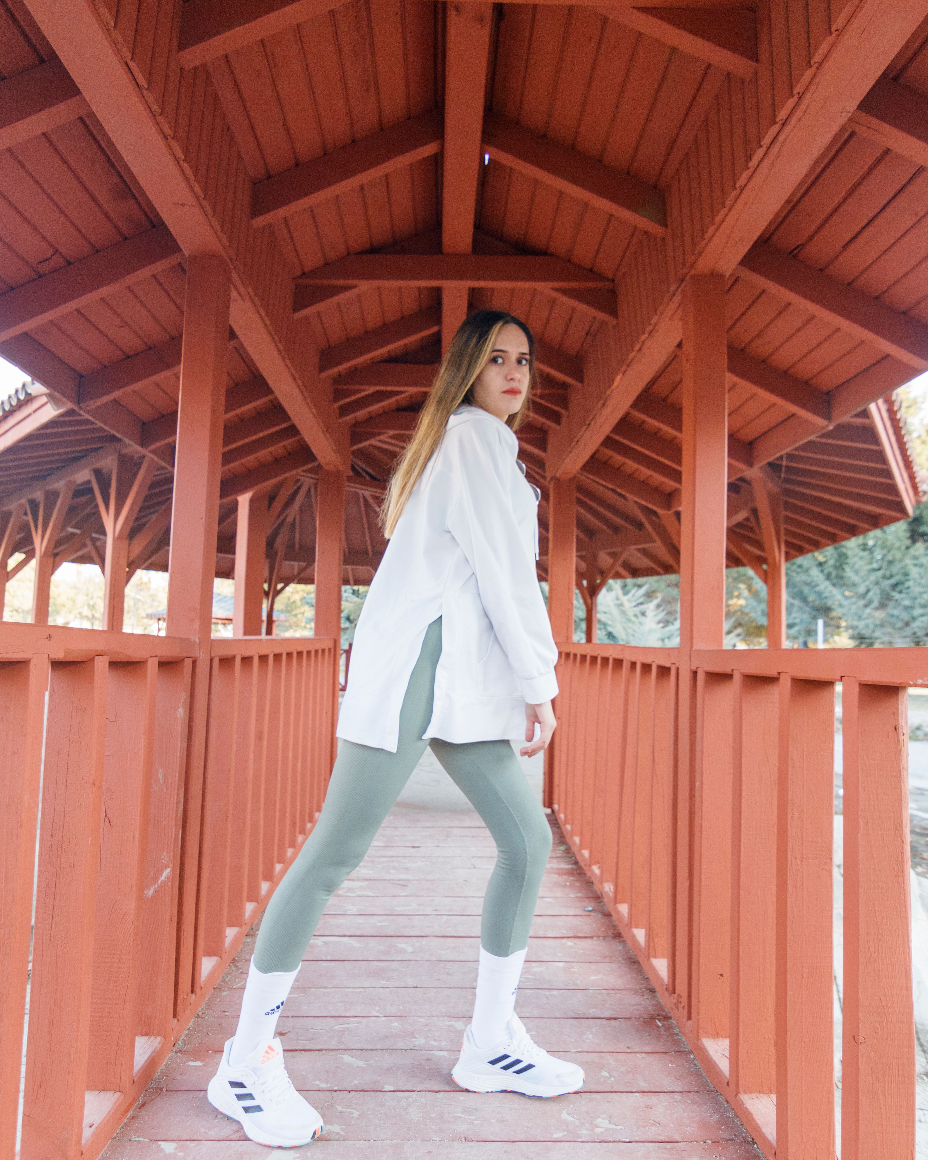 Jovem mulher em camisa branca e legging verde posa confiantemente em uma ponte de madeira ao ar livre.