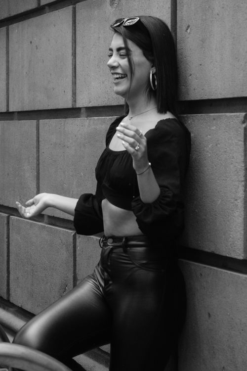 Free Black and white photo of a woman in leather pants laughing against a city wall. Stock Photo
