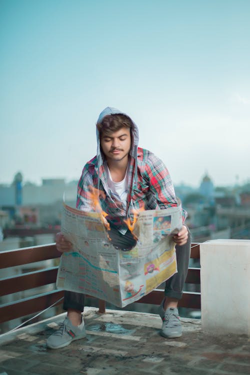 Man in Plaid Hoodie Long Sleeve Shirt Reading a Newspaper and Sitting on a Railing