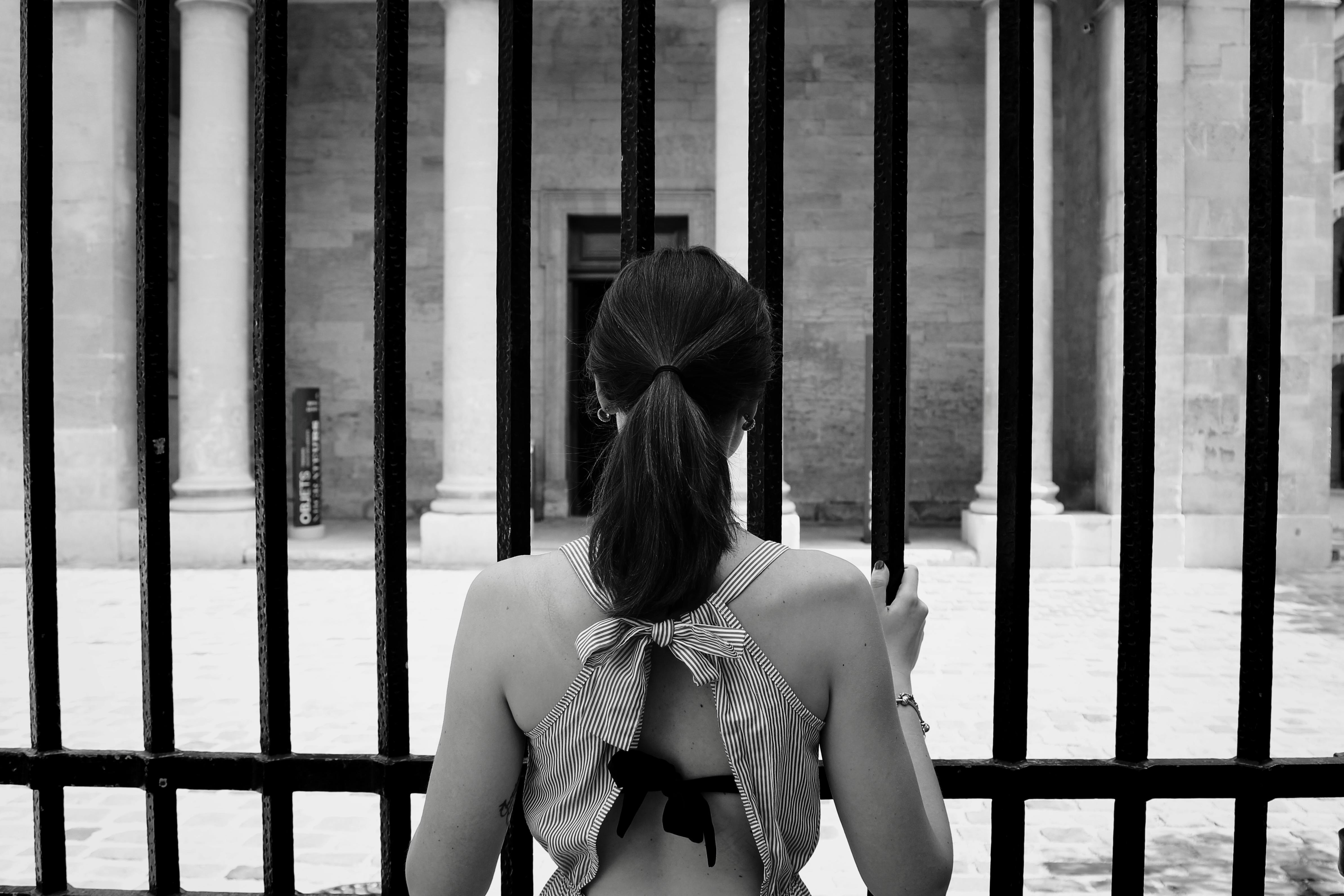 Back view of a woman looking through an iron fence at a historic building on a sunny day.
