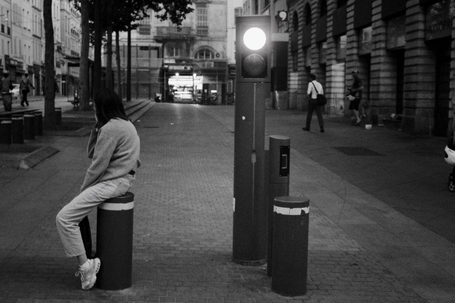 A woman in casual attire sitting on a bollard in a quiet urban city street at night.