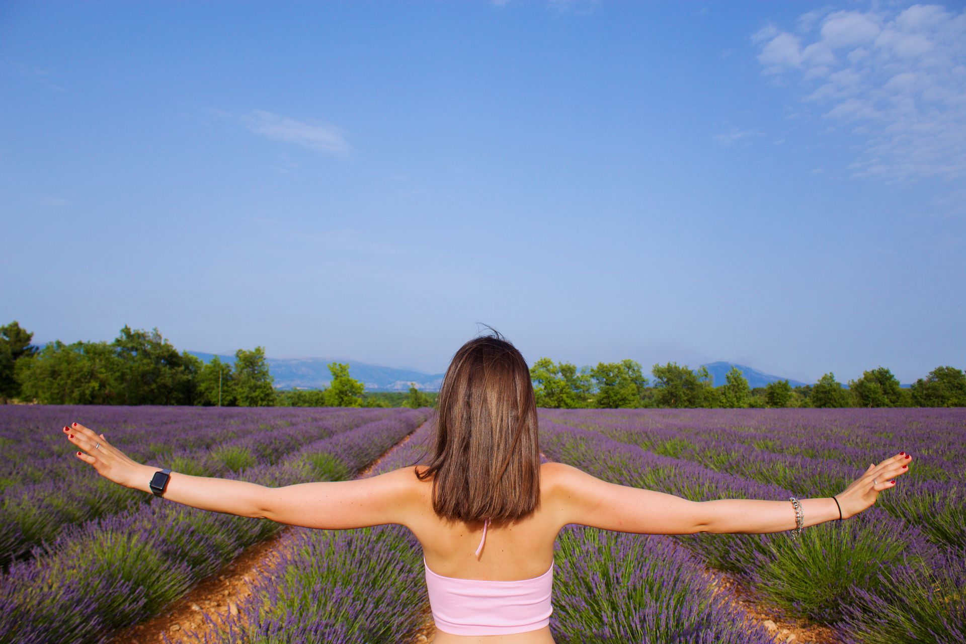 Back view of a woman with arms outstretched in a lavender field under a clear blue sky.