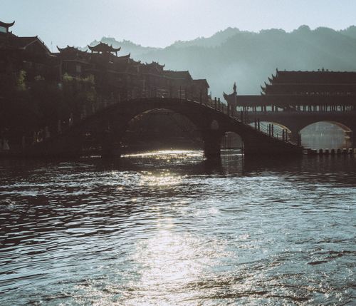 Free Traditional Chinese architecture bridge over a river, reflecting morning sunlight with misty hills in the background. Stock Photo
