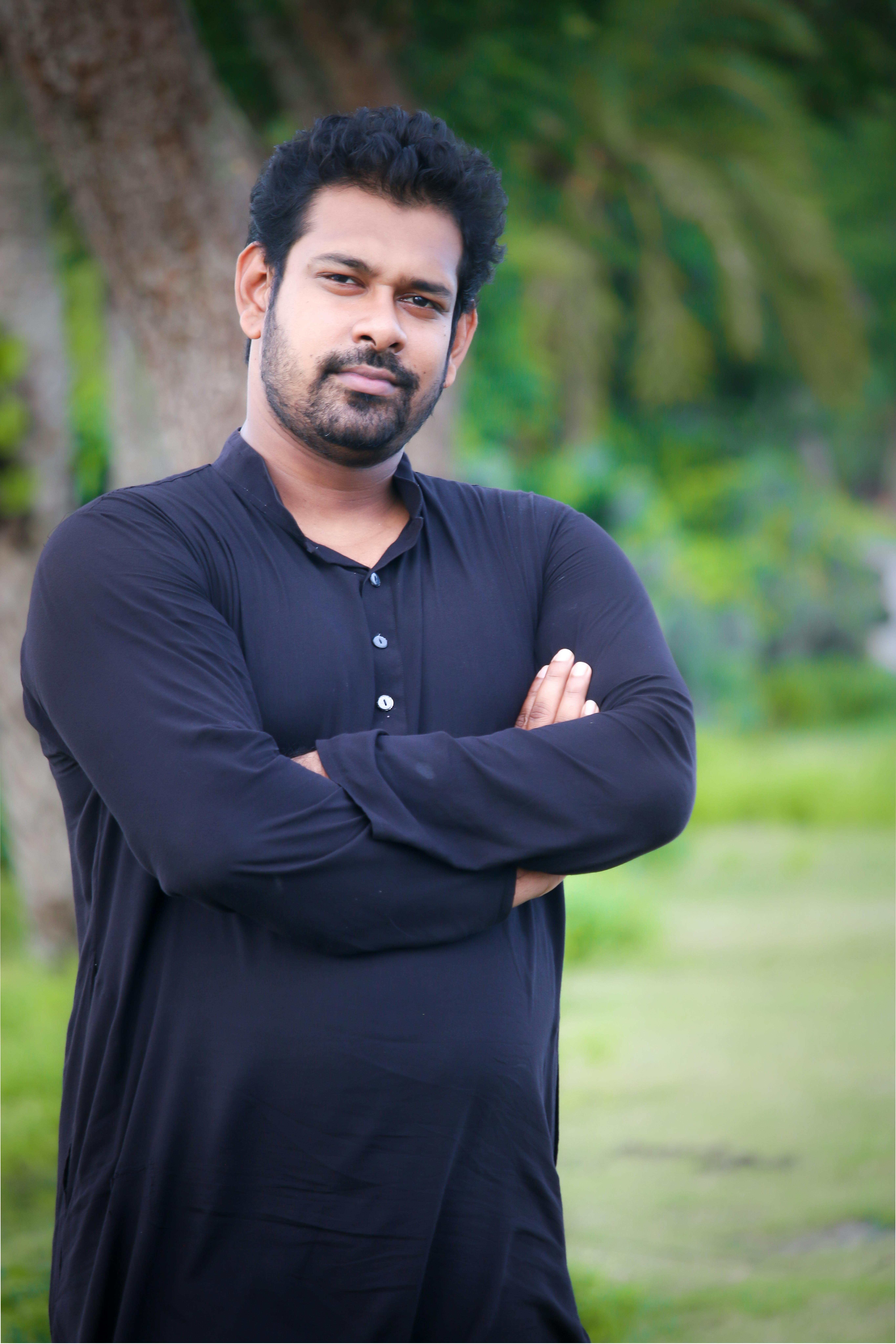 Portrait of a confident South Asian man in black, standing with arms crossed in a lush outdoor setting.